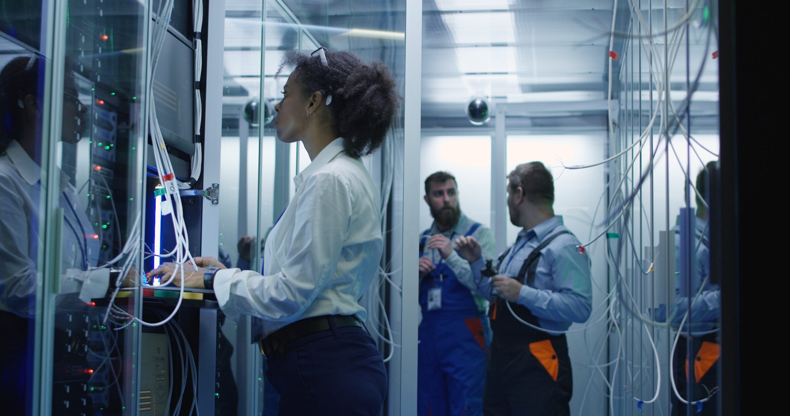 Technicians work in a data center, managing equipment and discussing tasks amid server racks and tangled wires, showcasing a collaborative technical environment.