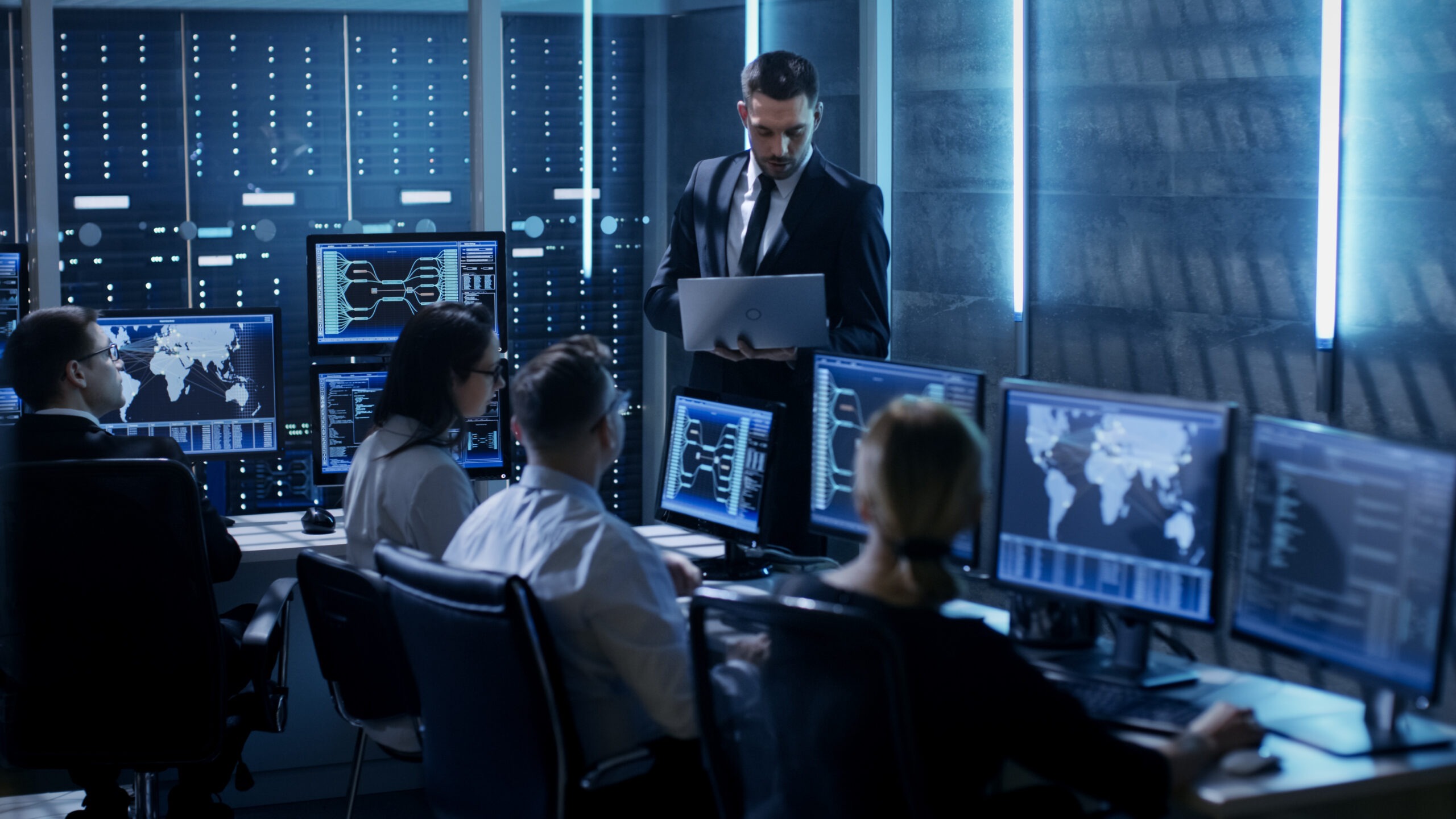 A group of professionals in business attire analyzes data on multiple screens in a high-tech server room. A man stands, holding a laptop, addressing the team.