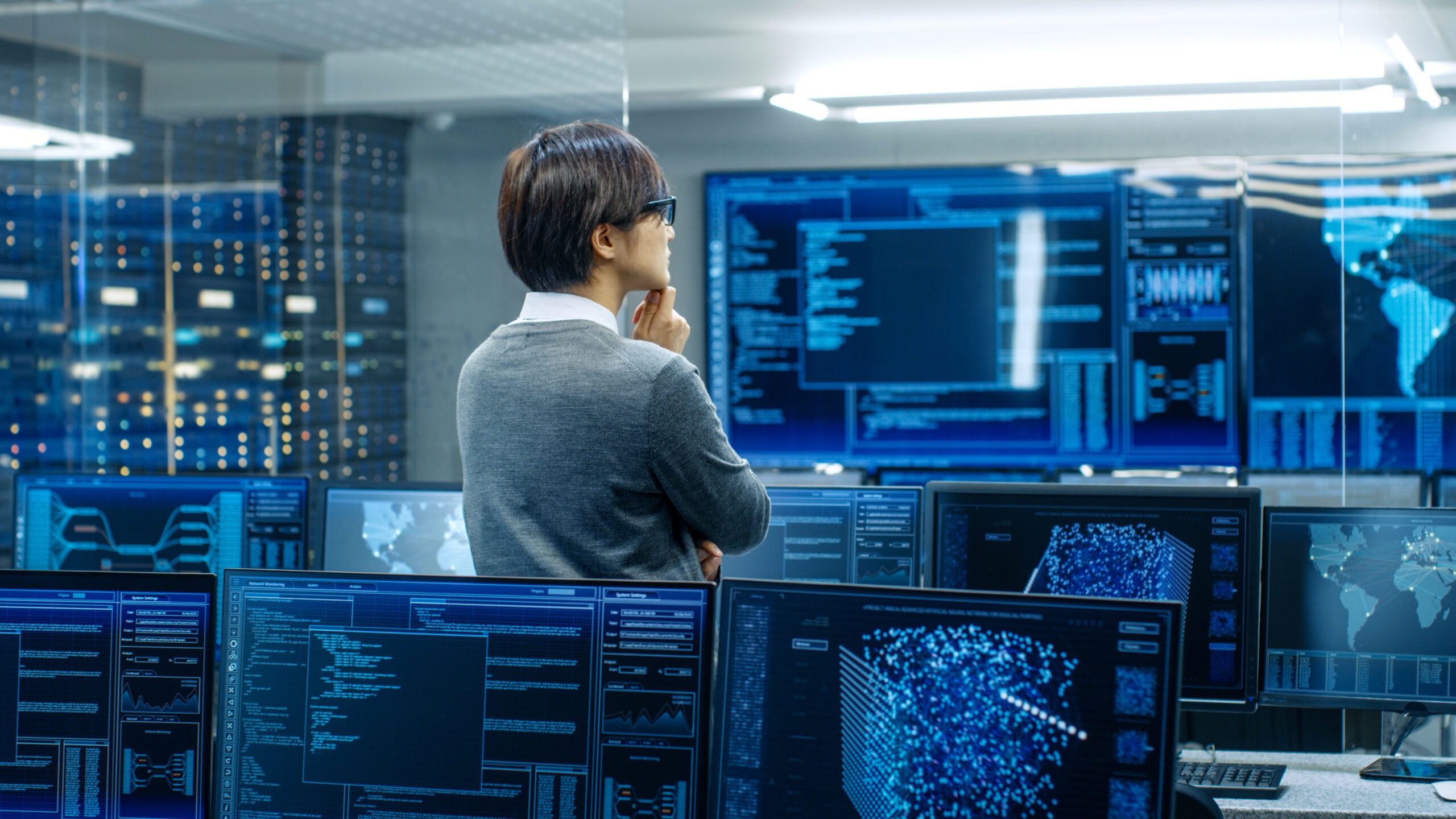 A data analyst stands pensively in a high-tech control room surrounded by multiple screens displaying coding and global network data.